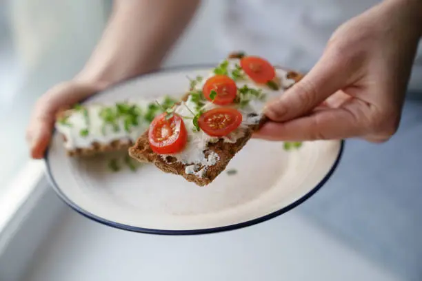 Woman hands holding plate with rye crisp bread with creamy vegetarian cheese tofu, cherry tomato and rucola micro greens. Healthy food, gluten free, diet concept.
