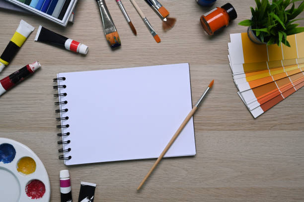 istock Artist workspace with empty notebook surrounded by color swatches, paintbrushes and other various equipment on wooden background. 1303103124