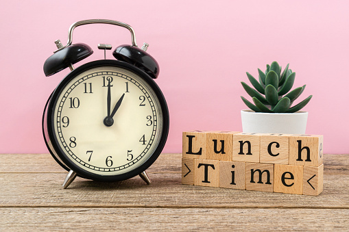 Black vintage alarm clock on a light pink background with a text lunch time on wooden block.