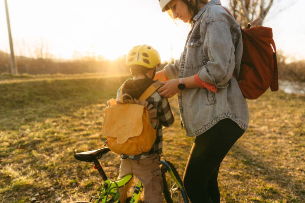 Photo of a little boy's commuting to school by bicycle. His mom's helping with his backpack.