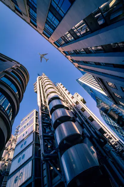 Highly detailed abstract wide angle view up towards the sky in the financial district of London City and its ultra modern contemporary buildings with unique architecture during a rare moment with commercial airplane passing / flying above the skyscrapers. Shot on Canon EOS R full frame with 14mm wide angle prime premium lens. Image is ideal for business travel concepts background.