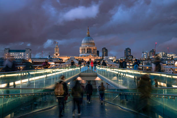 Motion blurred image providing a view of the busy Millennium Bridge with commuters and tourists on Thames Southbank towards the London city skyline with Saint Paul's Cathedral on a cloudy evening with moody sky. Shot with Canon EOS full frame system with extra wide lens. The image is ideal for horizontal background with plenty of copy space for your message.