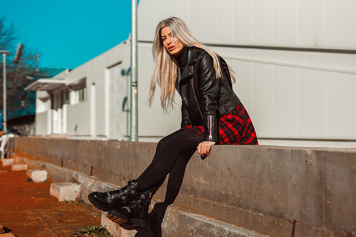 Fashionable young woman with long blond hair wearing a leather jacket, plaid skirt, and boots on a sunny winter day in the city