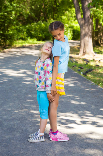 Two little sisters are measuring their height standing back to back in summer park Two cute little sisters are measuring their height standing back to back in summer park short stature stock pictures, royalty-free photos & images