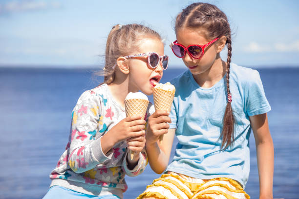 deux petites filles adorables mangent la crème glacée sur le bord de mer sur des vacances d’été. la petite fille goûte la glace de sa soeur - ice cream people food recreational pursuit photos et images de collection