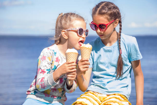 Two adorable little girls are eating ice cream on the seashore on summer vacation. Small girl is tasting her sister's ice cream Two adorable little girls wearing sunglasses are eating ice cream on the seashore on summer vacation. Small girl is tasting her sister's ice cream licking ice cream cone stock pictures, royalty-free photos & images