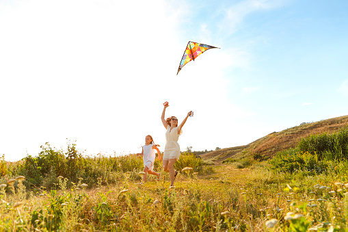 Carefree woman and teenage girl running along field and playing with kite during summer weekend
