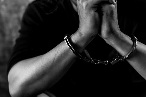 Criminal concept Bad guy was cuffed and under arrest by policeman and detain prisoner in jail Detainees get hopeless and stressed in dirty prison Young prisoner sit in dark room Black and white photo