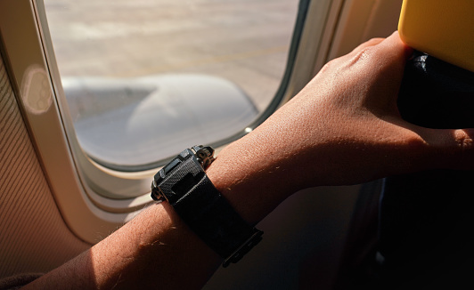 Sun shines on young man arm and plastic wristwatch near airplane window as he's waiting for takeoff