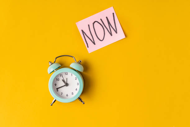 istock Alarm clock and a colored sticker with the inscription NOW on a grey background 1303133413