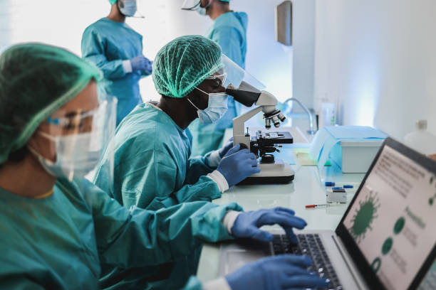 scienziato medico multirazziale in tuta hazmat che lavora con microscopio e computer portatile all'interno del moderno ospedale di laboratorio - focus volto dell'uomo africano - ricerca medica foto e immagini stock