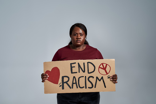 Sad plus size young african american woman in casual clothes looking at camera, holding End Racism banner in front of her, posing isolated over gray background. Social issues, protest concept