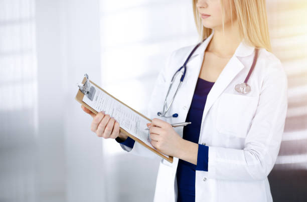 istock Unknown woman-doctor is writing some notes using a clipboard, while standing in the sunny cabinet in a clinic. Female physician with a stethoscope, close-up. Perfect medical service in a hospital. Medicine concept 1303136651