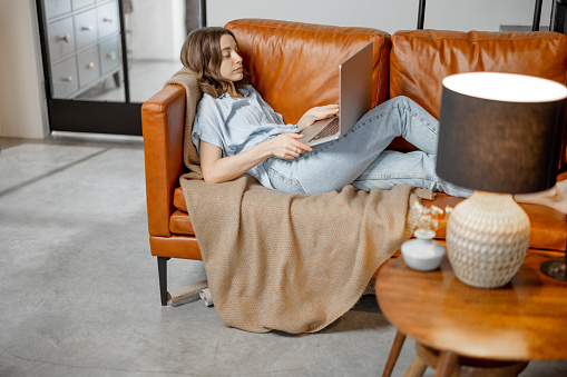 Young woman working with laptop while lying on foxy leather sofa. Digital nomad and work from home concept. High quality photo