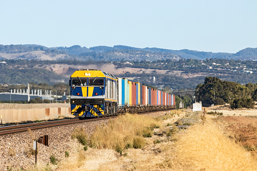 Twin diesel locomotives with container train passing industrial and farming area at speed with some summer heat haze. A range of tree-lined hills in the background. Logos and ID removed, paint scheme modified.