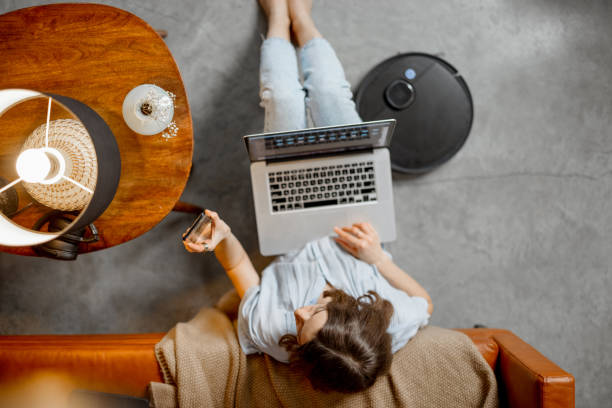 Woman working with laptop and Smartphone from home Woman working with laptop and looking on Smartphone from home while sitting on the floor near sofa. Robot vacuum cleaning at home . High quality photo home automation stock pictures, royalty-free photos & images