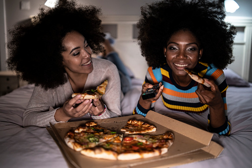 African women lying down on bed, eating pizza and watching TV.