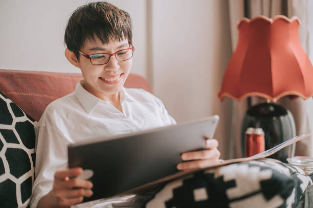 Asian chinese mid adult woman using digital tablet doing homework writing on notepad on sofa in living room