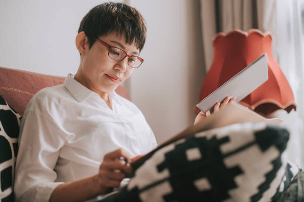 Asian chinese mid adult woman using digital tablet doing homework writing on notepad on sofa in living room Asian chinese mid adult woman using digital tablet doing homework writing on notepad on sofa in living room leanincollection stock pictures, royalty-free photos & images