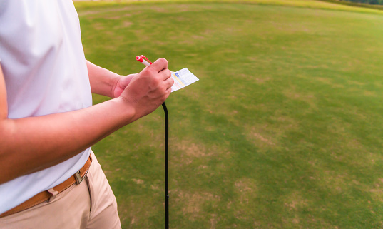 Asian male golfer using pen writing his golf score on scorecard while standing on green fairway.