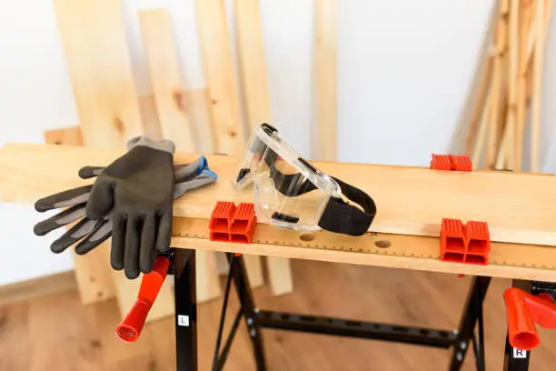 Workbench with protective glasses and safety gloves, without anyone. Inside a flat with wooden floors and white walls. It is daytime and there is natural light.