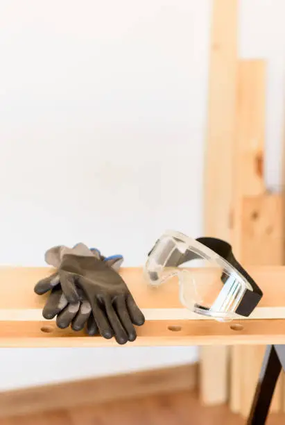 Workbench with protective glasses and safety gloves, without anyone. Inside a flat with wooden floors and white walls. It is daytime and there is natural light.