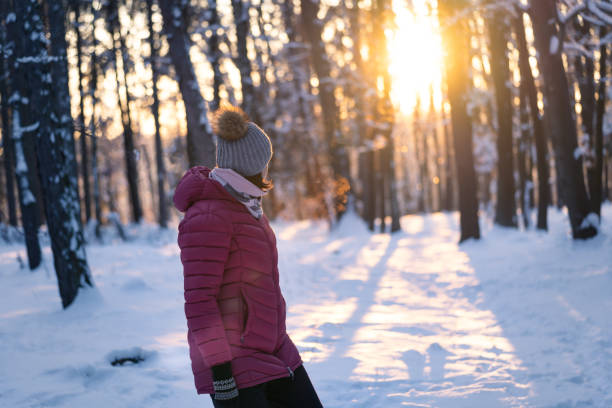 Middle aged caucasian woman soaking up the sun in forest in winter. Mature woman looking back through trees at beautiful sunset in winter. stock photo