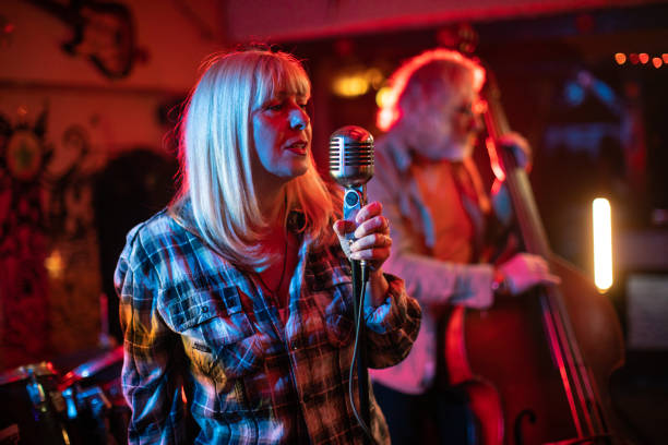 istock Senior woman with a microphone performing on stage with her band 1303157003