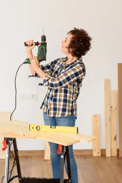 Caucasian woman with curly hair holds a drill in her hands, ready to make home improvements. She is standing in front of a workbench. She wears a plaid shirt and jeans. White background. Natural light. Background with wooden planks.