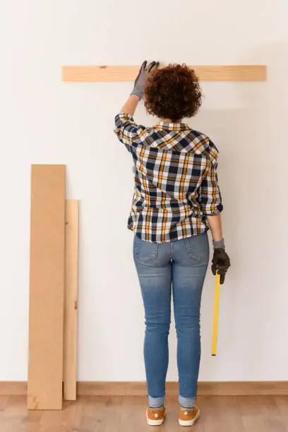 unrecognizable woman uses a level to accurately place a wooden plank on a white wall inside a flat with natural daylight.