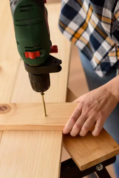 Close up of a drill drilling a screw into a piece of wood, on a wooden bench. Woman working on wooden bench. DIY enthusiast.