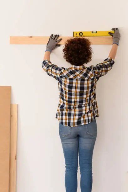 unrecognizable woman uses a level to accurately place a wooden plank on a white wall inside a flat with natural daylight.