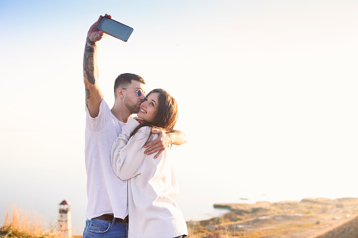 Positive young romantic couple in casual clothes hugging and kissing while taking selfie with smartphone on beach with lighthouse in background in sunny summer day