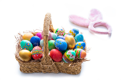 Easter eggs hand painted in a straw basket isolated on white with spring leaves and costume rabbit ears