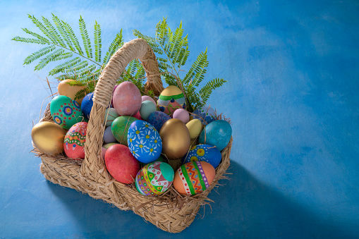 Easter eggs hand painted in a straw basket on blue background copy space with spring grass leaves