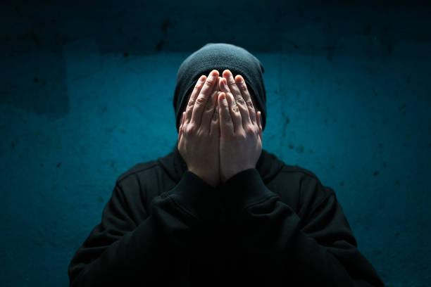 Color image depicting an adult man wearing a black beanie hat in an underground subway tunnel. His expression is one of despair, sadness and depression. His hands are covering his face in a gesture of bleak desperation and hopelessness. Room for copy space.