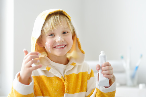 Cute blonde caucasian boy applying cream on his face in bathroom in the morning. Kids care cosmetics. Hygiene for little child.