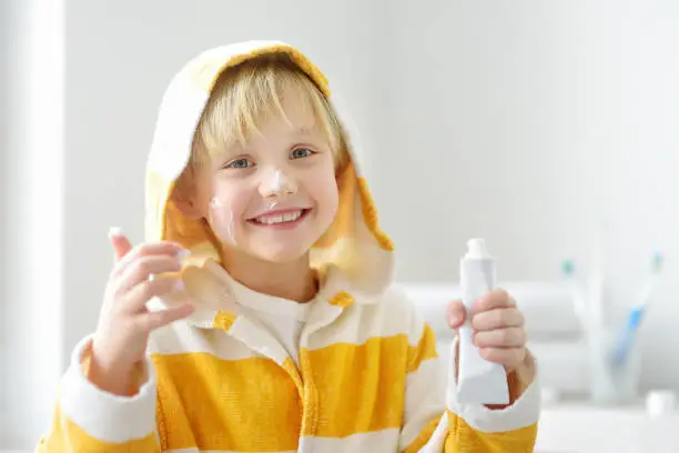 Photo of Cute blonde caucasian boy applying cream on his face in bathroom in the morning. Kids care cosmetics. Hygiene for child.