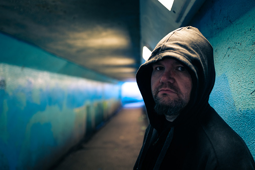 Color image depicting an adult man in his 30s in an underground subway tunnel. He is wearing a hooded sweater with the hood pulled up over his head. His expression is one of despair, sadness and depression. Focus is on the man in the foreground while we can see the light at the end of the dark and moody tunnel in the background. Room for copy space.