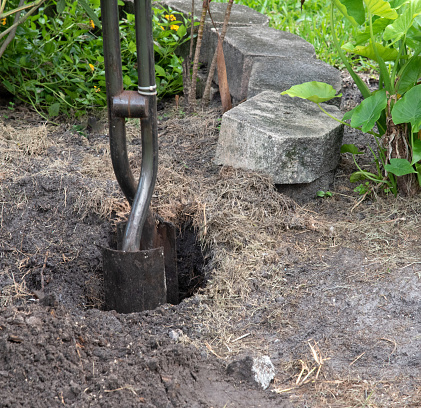 Steel post hole digger with metal shovel-like blades is being used to  create a deep hole for a fence post with concrete bricks and green plants in the background.