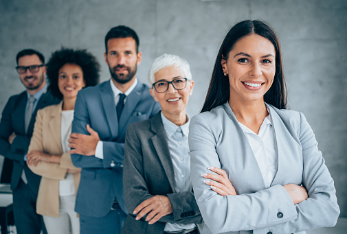 Portrait of beautiful smiling businesswoman with her colleagues. Multi-ethnic group of business persons standing in a row in modern office. Successful team leader and her team in background.