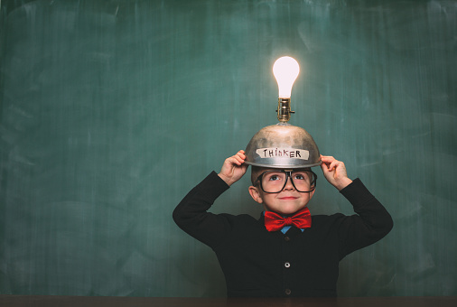 A young nerd boy dressed in retro 1980s attire, with bow tie and eyeglasses, wears a light bulb idea invention machine to help him think of the next big idea.