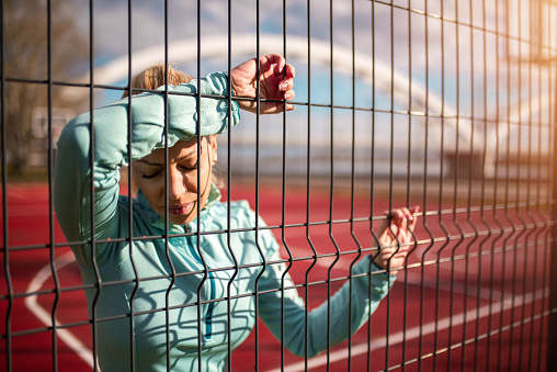Depressed and stuck mid adult woman standing behind bars. She is a sporty woman standing on a basketball court feeling stuck in life. Concept photo.