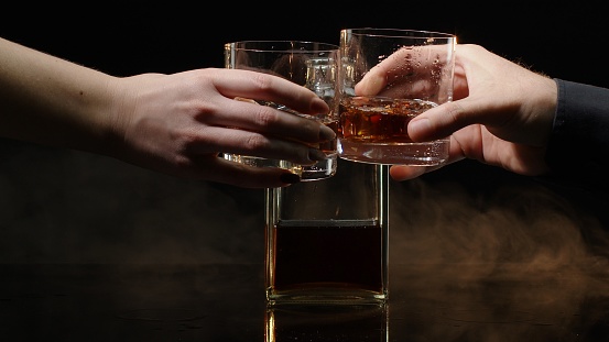 Two hands with glasses of cognac whiskey with ice cubes making cheers on black background. Celebration of business success, Christmas, anniversary, birthday. Raising toast with glasses of bourbon, rum