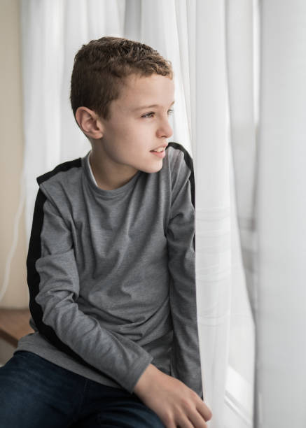 istock Young boy stuck indoors sat on windowsill looking through curtain window outside with cheerful smile on bright day 1303207938