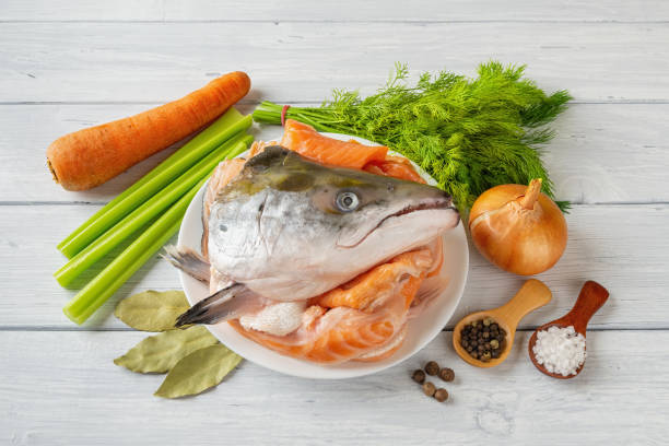 魚のストックを調理するための原料:新鮮なサーモンの頭と骨、野菜、塩、コショウを白い木のテーブルの上に。赤い魚のスープブイヨンのレシピ。自家製シーフードの健康的な食事。 - pink pepper ストックフォトと画像