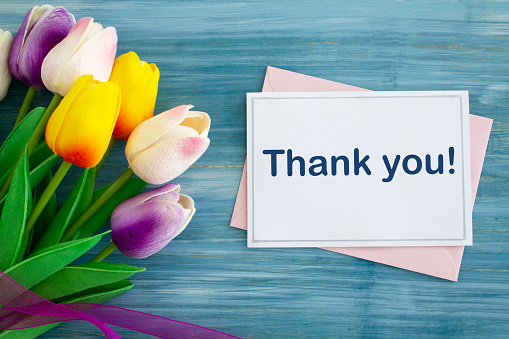 A thank you card with brightly coloured tulips and ribbon on blue rustic wood.\nShot from above.