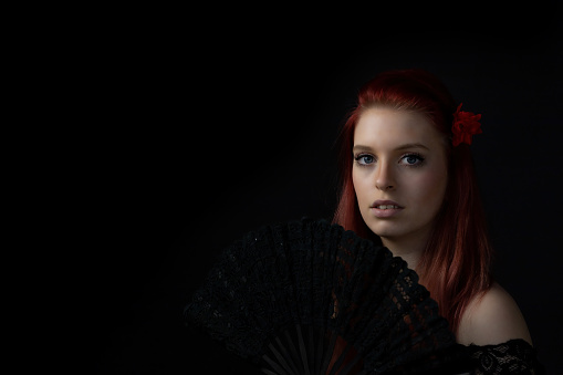 Low key portrait of beautiful young woman with fan looking at the camera. Horizontally.