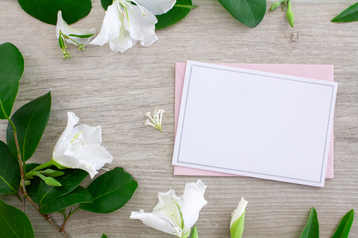 A blank card with a pink envelope with fresh white flowers and leaves on a rustic table. 
Shot from above.