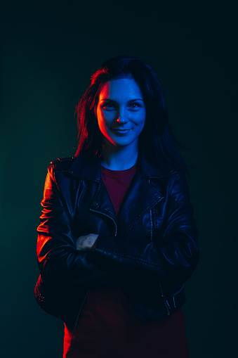 Night female portrait. Neon light. Positive attitude. Confident happy woman standing with folded arms looking at camera smiling in red blue glow isolated on dark teal background.